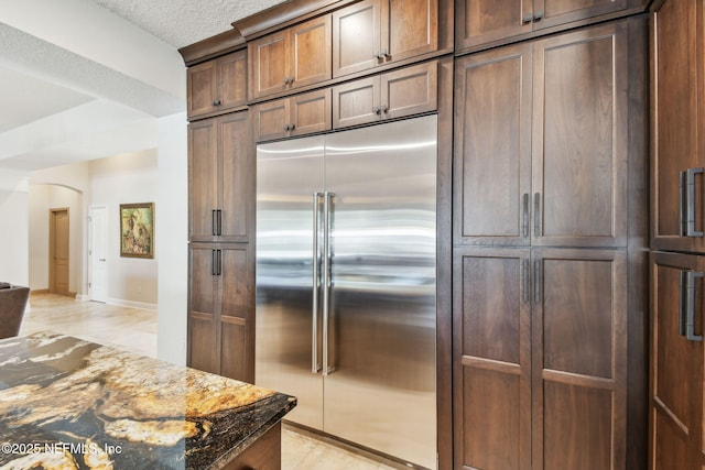 kitchen with stainless steel built in refrigerator, a textured ceiling, dark brown cabinetry, and dark stone counters