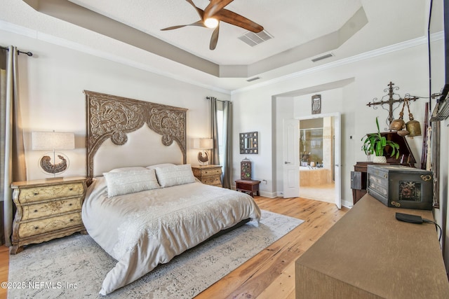 bedroom featuring light wood-type flooring, a raised ceiling, ceiling fan, crown molding, and connected bathroom