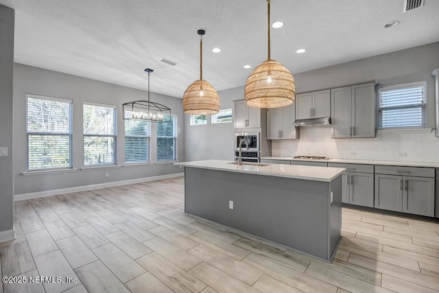 kitchen with gray cabinets, decorative light fixtures, stainless steel gas stovetop, an island with sink, and sink