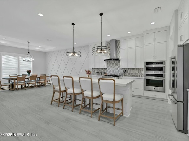 kitchen with a center island with sink, wall chimney range hood, hanging light fixtures, appliances with stainless steel finishes, and white cabinetry