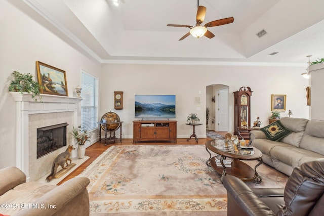 living room with light hardwood / wood-style flooring, ornamental molding, a tray ceiling, a fireplace, and ceiling fan