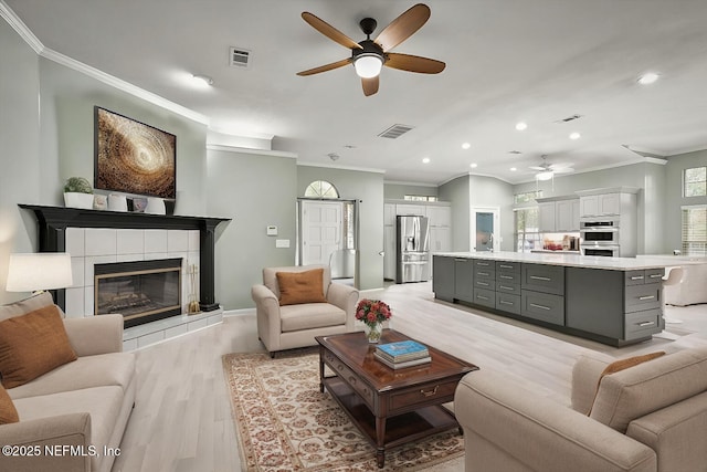 living room featuring ceiling fan, a tiled fireplace, crown molding, and light hardwood / wood-style flooring