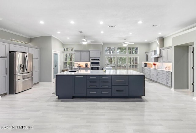 kitchen with sink, wall chimney range hood, an island with sink, gray cabinets, and appliances with stainless steel finishes