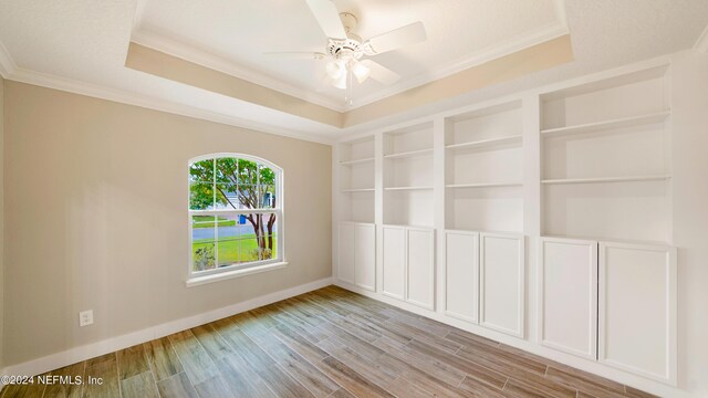 spare room with ceiling fan, built in features, crown molding, a tray ceiling, and light wood-type flooring