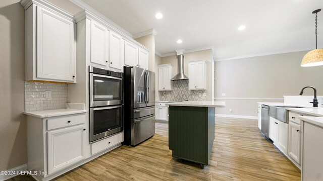 kitchen with white cabinetry, hanging light fixtures, stainless steel appliances, wall chimney range hood, and ornamental molding