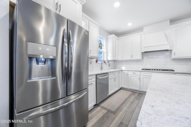 kitchen with light stone counters, custom exhaust hood, stainless steel appliances, wood-type flooring, and white cabinetry