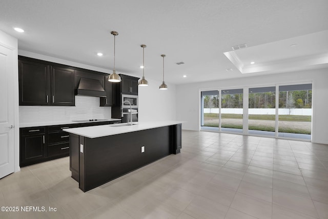 kitchen with premium range hood, decorative light fixtures, sink, black electric stovetop, and a center island with sink