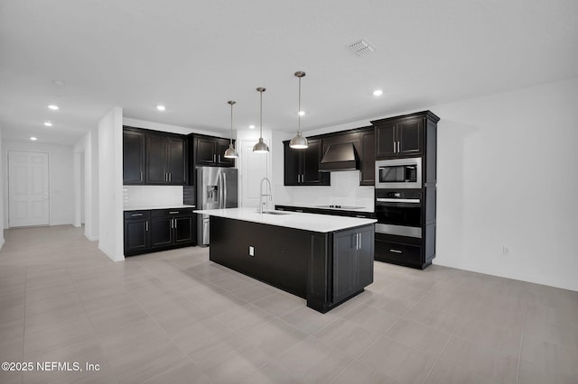 kitchen with appliances with stainless steel finishes, decorative light fixtures, an island with sink, sink, and wall chimney exhaust hood