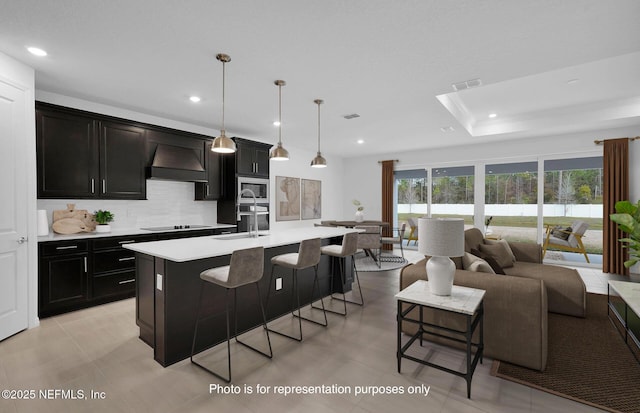 kitchen with sink, hanging light fixtures, custom range hood, a center island with sink, and black electric cooktop