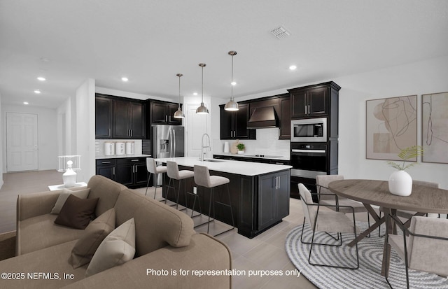 kitchen featuring sink, custom exhaust hood, decorative light fixtures, appliances with stainless steel finishes, and a kitchen island with sink