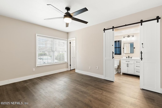 unfurnished bedroom with dark wood-style flooring, ensuite bathroom, a ceiling fan, a barn door, and baseboards