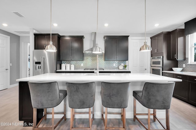 kitchen with pendant lighting, a center island with sink, light hardwood / wood-style flooring, wall chimney exhaust hood, and appliances with stainless steel finishes