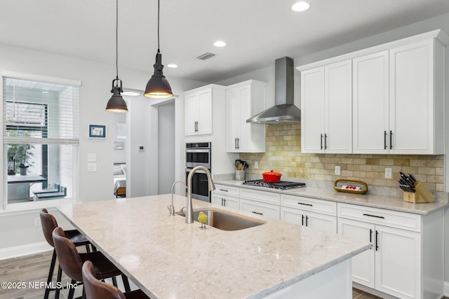 kitchen with sink, hanging light fixtures, stainless steel appliances, an island with sink, and wall chimney exhaust hood