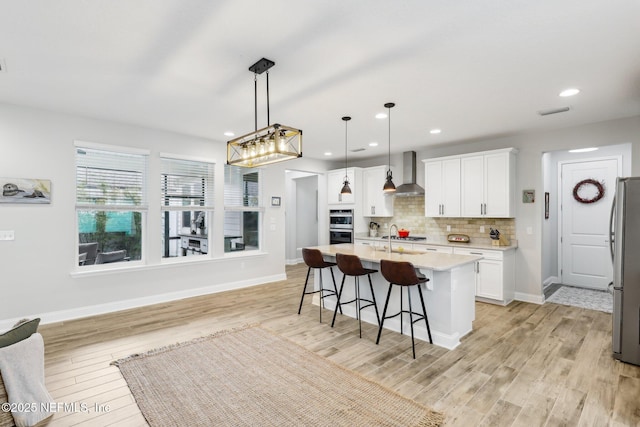 kitchen featuring pendant lighting, white cabinetry, a kitchen island with sink, stainless steel appliances, and wall chimney exhaust hood