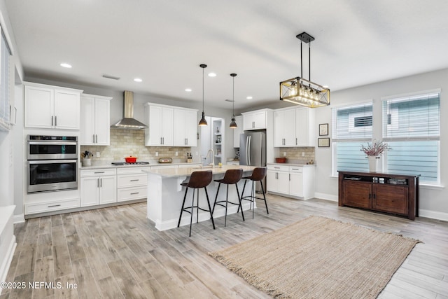 kitchen featuring stainless steel appliances, a kitchen island with sink, wall chimney range hood, and white cabinets