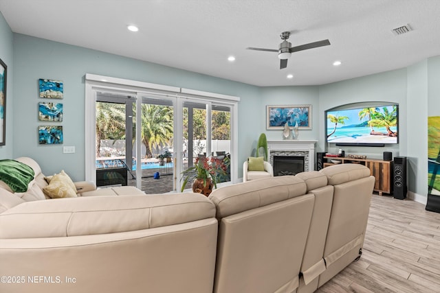 living room featuring ceiling fan, a stone fireplace, a textured ceiling, and light hardwood / wood-style flooring