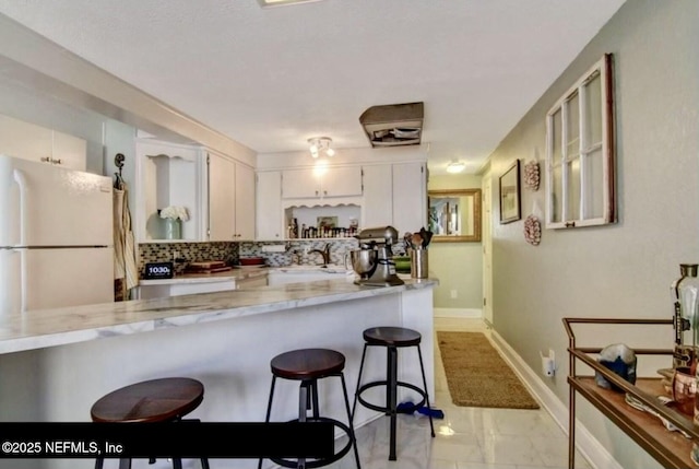 kitchen featuring backsplash, white cabinets, white refrigerator, light stone countertops, and a breakfast bar area