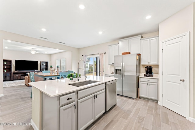 kitchen with light stone counters, stainless steel appliances, a center island with sink, ceiling fan, and sink