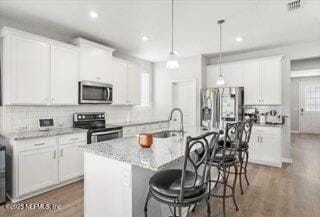 kitchen with white cabinetry, stainless steel appliances, light stone counters, pendant lighting, and a kitchen island with sink