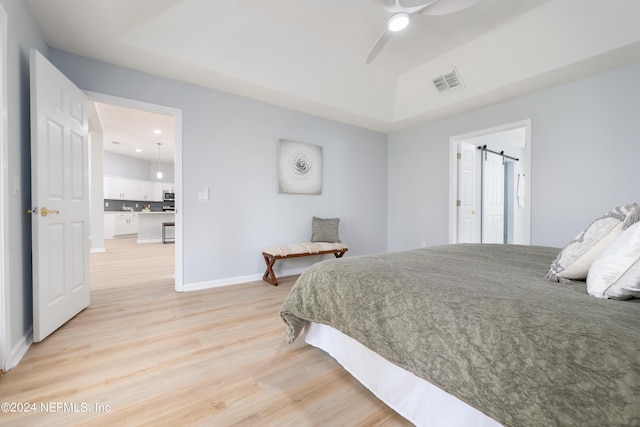 bedroom with a barn door, light hardwood / wood-style floors, ceiling fan, and a tray ceiling