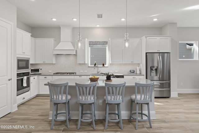 kitchen featuring light wood-style flooring, a kitchen island, appliances with stainless steel finishes, premium range hood, and backsplash