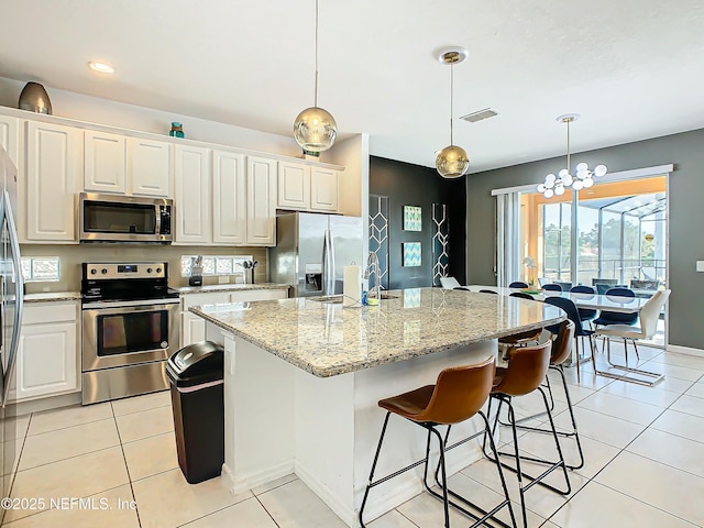 kitchen with an island with sink, decorative light fixtures, light tile patterned flooring, light stone counters, and stainless steel appliances