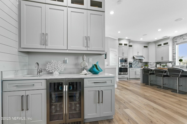 bar featuring under cabinet range hood, beverage cooler, stainless steel appliances, a sink, and light wood finished floors