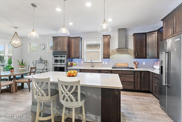 kitchen with tasteful backsplash, wall chimney range hood, a center island, hanging light fixtures, and appliances with stainless steel finishes