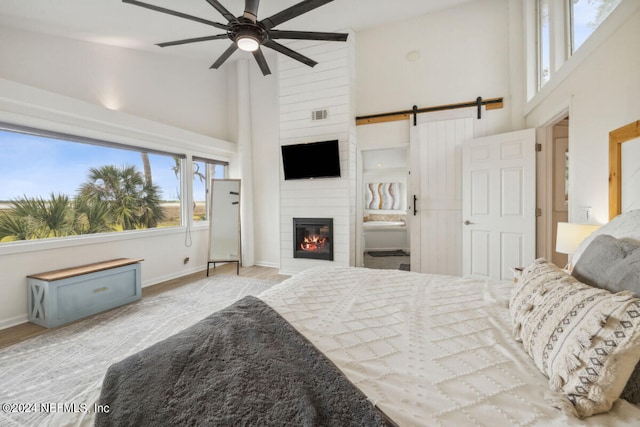 bedroom with a large fireplace, ceiling fan, a barn door, a high ceiling, and connected bathroom