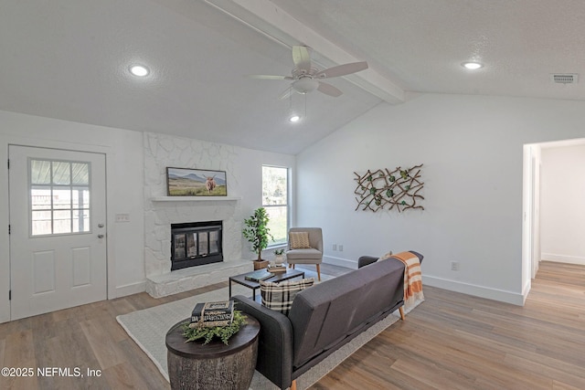 living room featuring a textured ceiling, ceiling fan, vaulted ceiling with beams, light hardwood / wood-style floors, and a stone fireplace