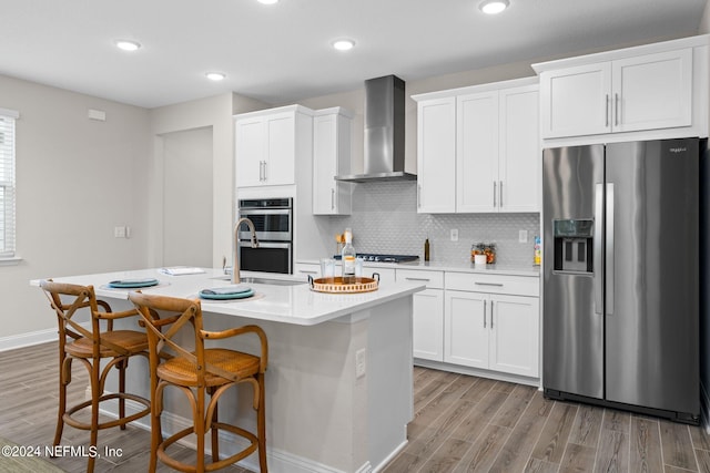 kitchen featuring a kitchen island with sink, white cabinets, stainless steel appliances, and wall chimney range hood