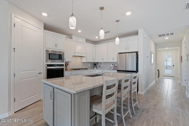 kitchen with stainless steel appliances, sink, white cabinetry, light stone counters, and a center island with sink