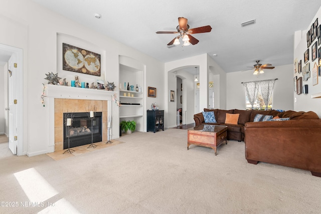 living area featuring visible vents, arched walkways, a tiled fireplace, light colored carpet, and built in shelves