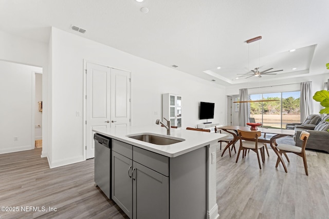 kitchen with gray cabinetry, dishwasher, sink, a raised ceiling, and a center island with sink