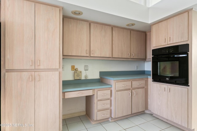 kitchen with light brown cabinetry, built in desk, black oven, and light tile patterned flooring