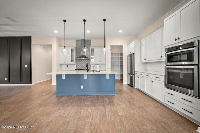 kitchen with a kitchen island with sink, white cabinets, hanging light fixtures, wall chimney exhaust hood, and stainless steel appliances