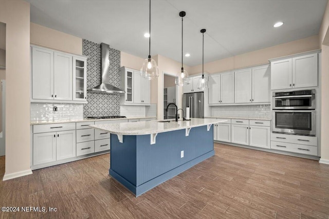 kitchen with appliances with stainless steel finishes, wall chimney exhaust hood, a center island with sink, decorative light fixtures, and white cabinets