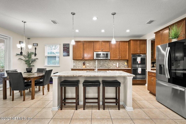 kitchen with tasteful backsplash, appliances with stainless steel finishes, a center island with sink, and decorative light fixtures