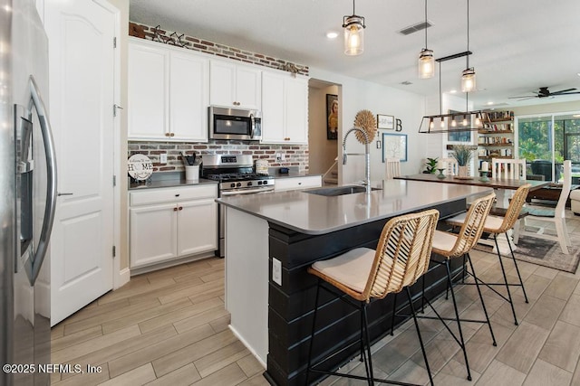 kitchen with stainless steel appliances, ceiling fan, decorative light fixtures, white cabinets, and an island with sink