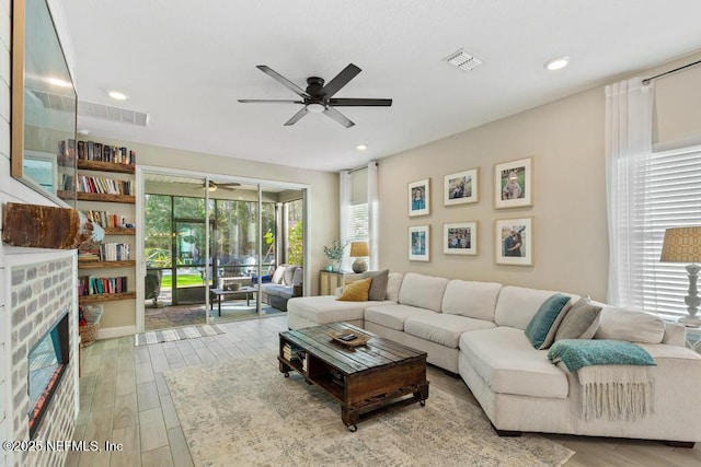 living area featuring a brick fireplace, ceiling fan, visible vents, and wood finished floors