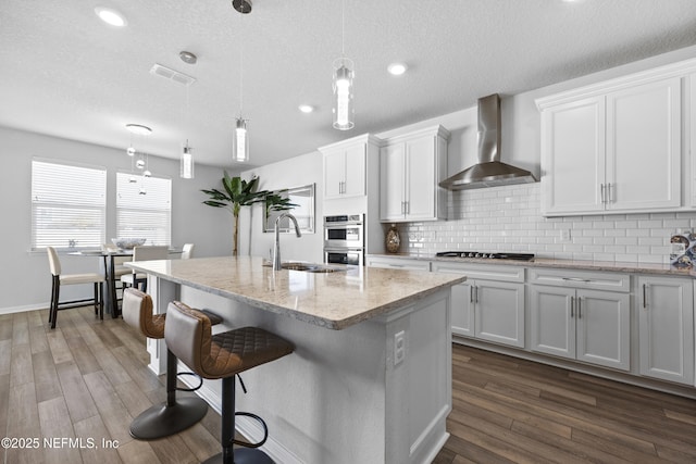 kitchen featuring pendant lighting, an island with sink, white cabinets, and wall chimney range hood