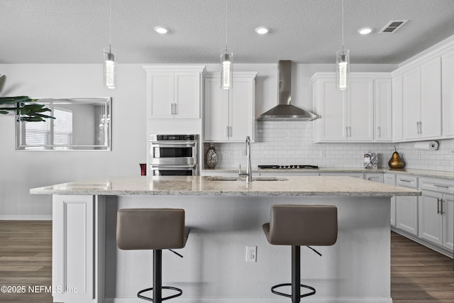 kitchen with decorative light fixtures, a kitchen island with sink, white cabinetry, and wall chimney range hood