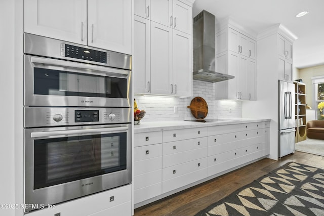 kitchen with wall chimney range hood, stainless steel appliances, white cabinets, dark hardwood / wood-style flooring, and decorative backsplash