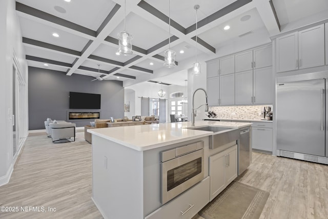 kitchen with built in appliances, an island with sink, backsplash, hanging light fixtures, and light hardwood / wood-style flooring