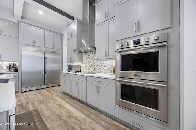 kitchen featuring appliances with stainless steel finishes, wall chimney exhaust hood, tasteful backsplash, light hardwood / wood-style floors, and light stone counters