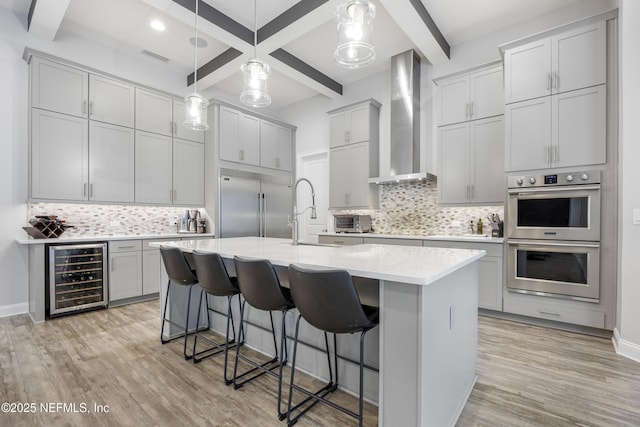 kitchen featuring wine cooler, appliances with stainless steel finishes, light wood-type flooring, an island with sink, and wall chimney exhaust hood