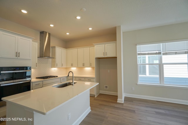 kitchen with white cabinets, a center island with sink, wall chimney range hood, and sink