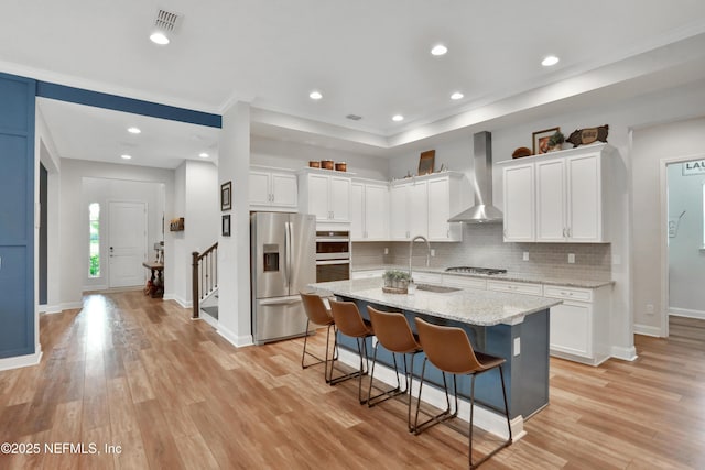 kitchen with stainless steel appliances, a center island with sink, wall chimney range hood, and white cabinetry