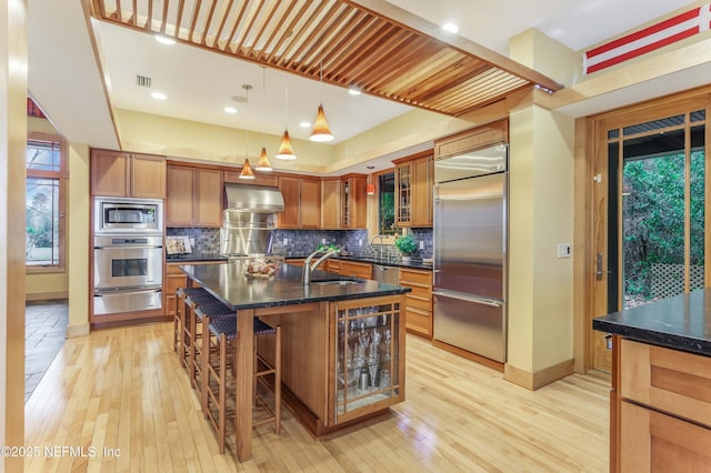 kitchen with sink, hanging light fixtures, built in appliances, a center island with sink, and wall chimney range hood