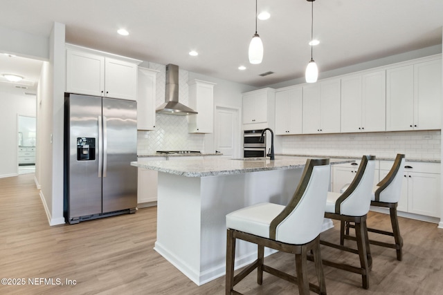 kitchen with hanging light fixtures, a kitchen island with sink, appliances with stainless steel finishes, white cabinets, and wall chimney exhaust hood